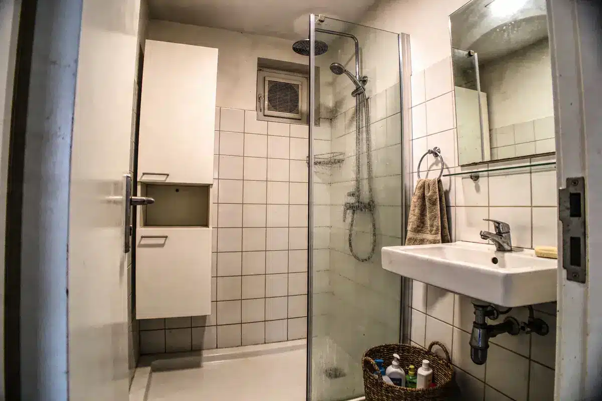 Petite salle de bain carrelée, douche vitrée, lavabo mural, étagère, miroir, panier avec produits de toilette.