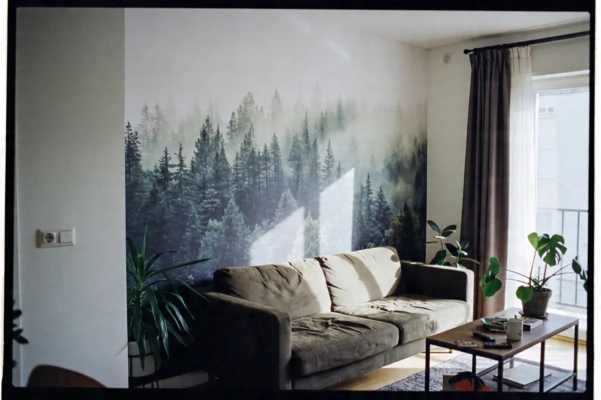 Salon avec canapé, mur décoré de forêt, plantes vertes, table basse en bois, lumière naturelle par fenêtre.