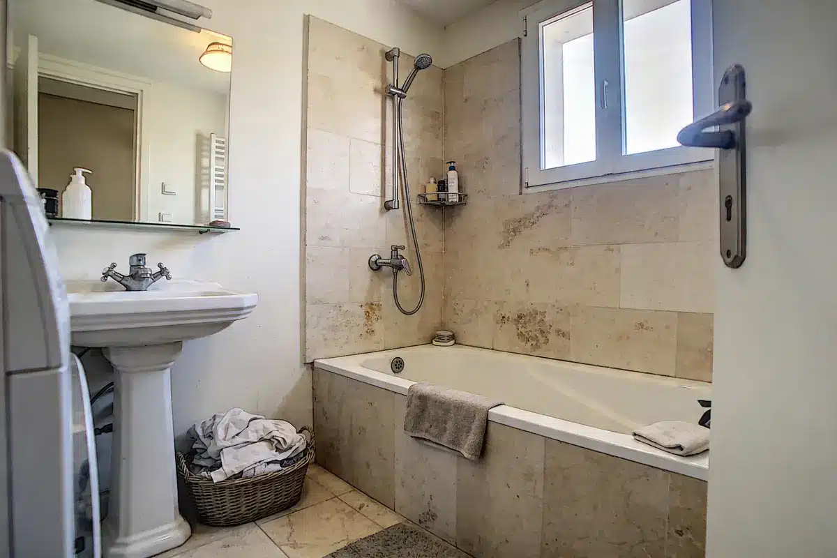 Salle de bain avec baignoire, lavabo, fenêtre, miroir, panier linge et produits de toilette.
