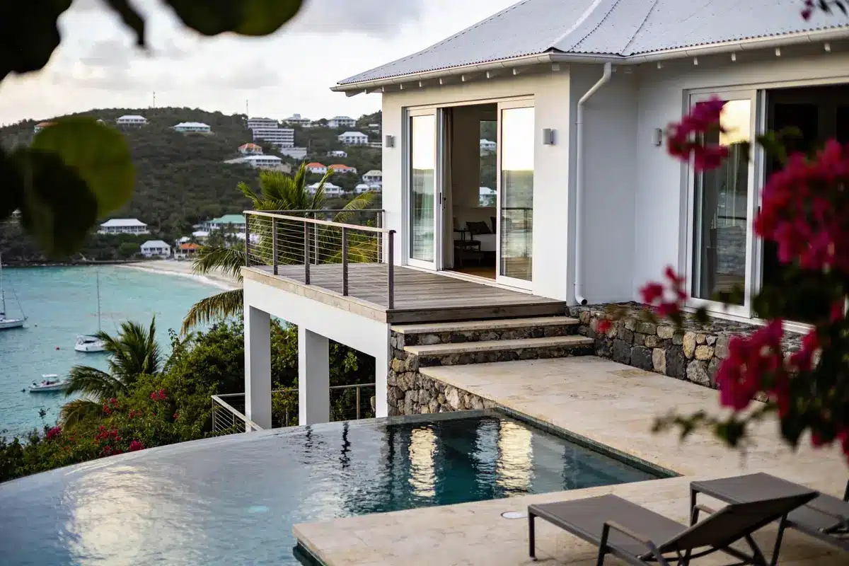 Maison moderne avec piscine à débordement, vue mer et collines, entourée de végétation tropicale.