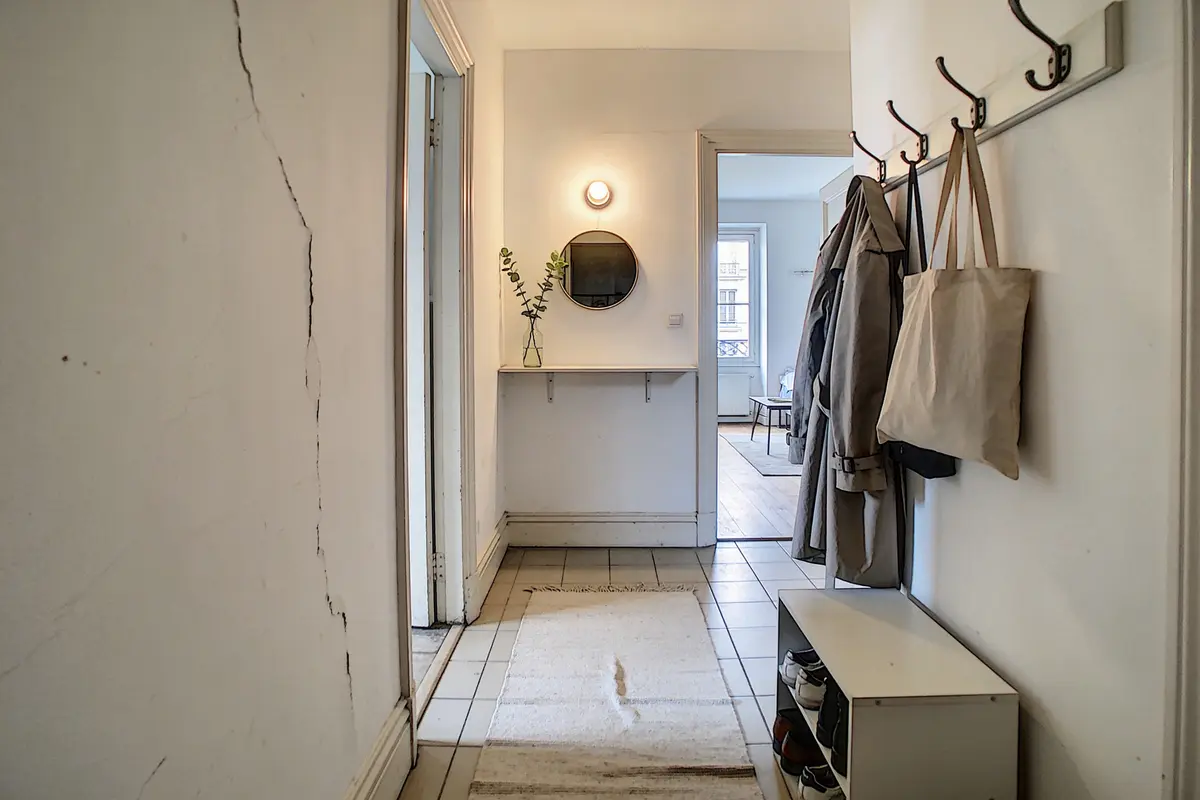Couloir intérieur lumineux avec porte-manteaux, sac en toile, tapis beige et miroir rond sur mur blanc.